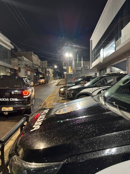 Imagem noturna de uma rua com viaturas policiais estacionadas. A cena é iluminada por luzes artificiais, revelando prédios e um ambiente urbano.