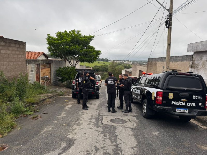 Agentes da Polícia Civil conversando em uma rua, com viaturas estacionadas ao fundo, cercados por árvores e casas.