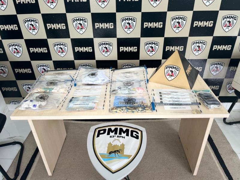 Mesa com diversos itens organizados em sacos plásticos, incluindo materiais e documentos, com fundo de parede estampada com logotipos da PMMG.