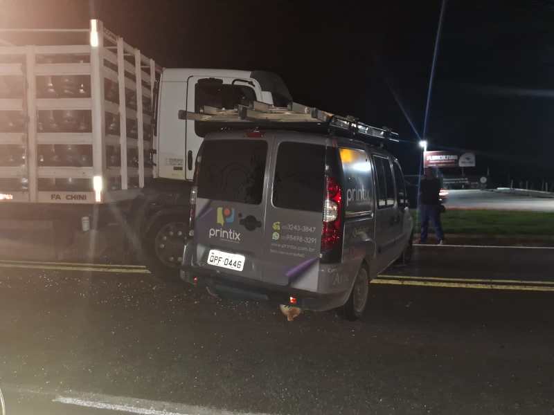 Uma van cinza da empresa Printix estacionada à noite, ao lado de um caminhão branco carregado. A cena é iluminada por luzes ao fundo.