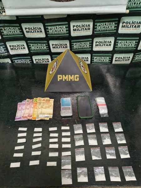Materiais apreendidos, incluindo dinheiro em notas, dois celulares e pacotes com substâncias suspeitas, dispostos em uma superfície escura, com fundo de placas da Polícia Militar.