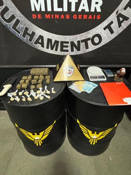 Drogas e dinheiro dispostos sobre barris, em um ambiente policial, com fundo de banner institucional.