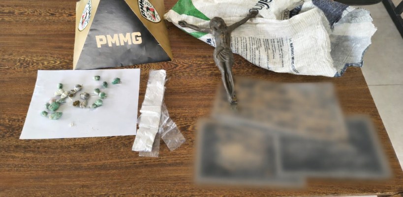 Diversos itens sobre uma mesa, incluindo um crucifixo, pequenos pacotes com substâncias, e um papel com logo da PMMG.