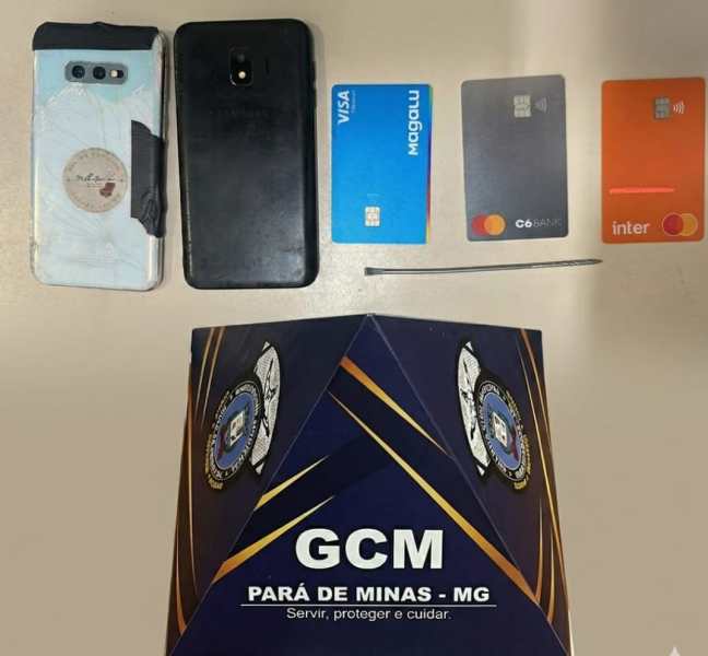 Um celular, vários cartões de crédito e um objeto metálico sobre uma mesa, ao lado de um folder da GCM de Pará de Minas, MG.