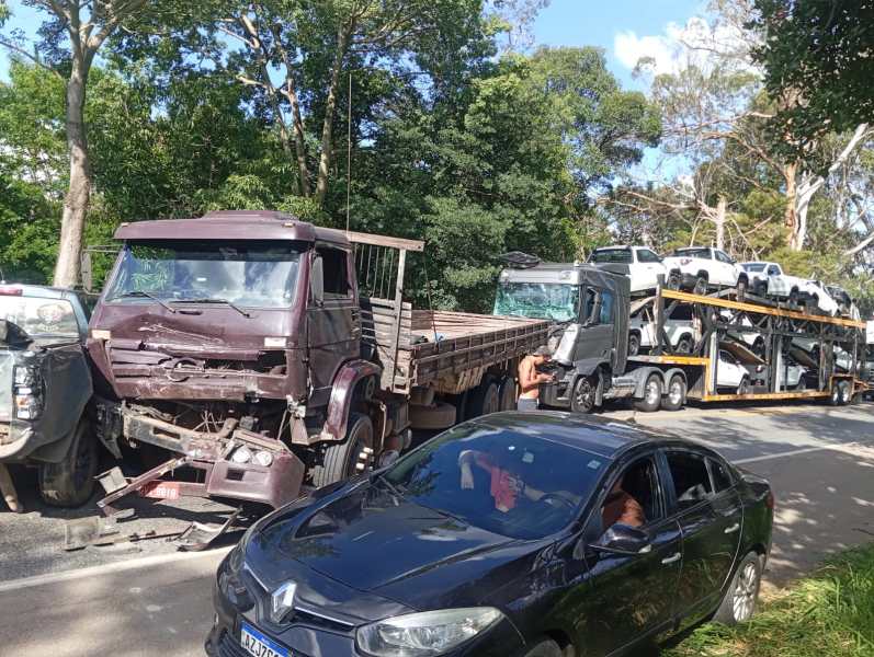 Acidente de trânsito envolvendo vários caminhões e um carro na estrada, com um caminhão carregando veículos novos e outro danificado.
