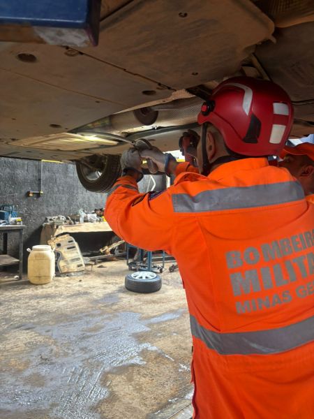 Um bombeiro militar trabalha embaixo de um veículo, utilizando uma ferramenta. Ele veste um traje laranja com refletores e um capacete vermelho. Ao fundo, há equipamentos e ferramentas em um ambiente de oficina.