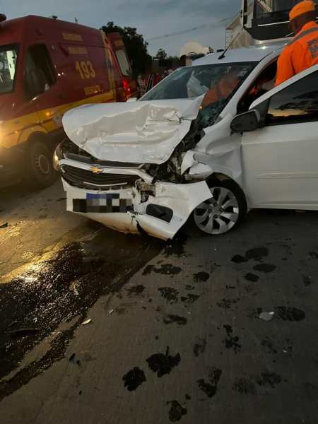 Carro branco amassado na frente após um acidente com a presença de veículos de emergência ao fundo.