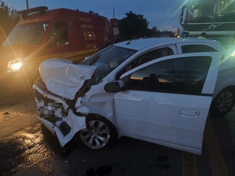 Veículo branco danificado após um acidente, com a parte frontal bastante amassada. Ao fundo, um caminhão de emergência e um outro veículo visíveis.