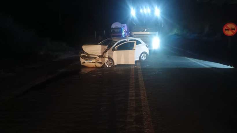 Carro branco com danos frontais parado em uma estrada à noite, com caminhão iluminado ao fundo e sinalização proibida.