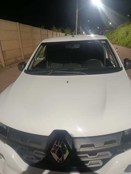 Imagem frontal de um carro branco da Renault, com um adesivo da Uber no para-brisa, à noite em uma via iluminada.