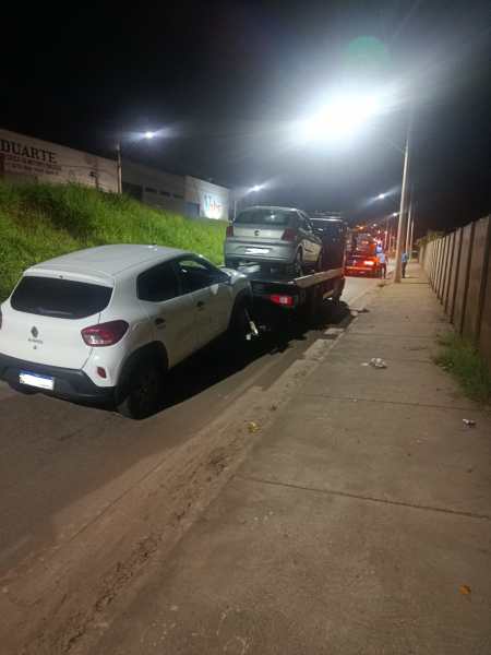 Um carro branco sendo rebocado à noite, com uma caminhonete de reboque transportando outros veículos em uma rua iluminada.