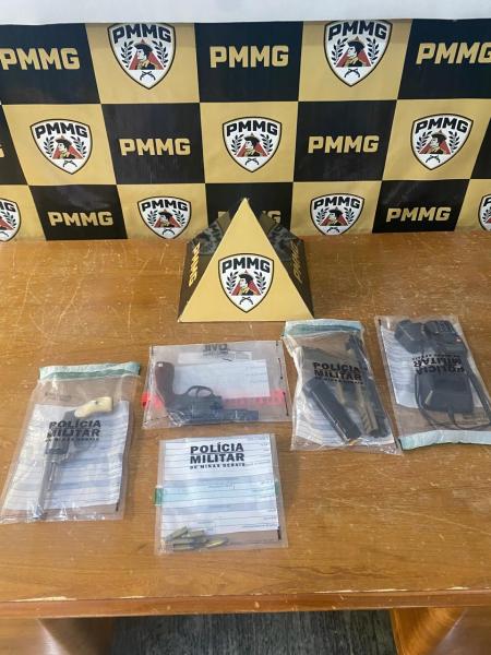 Material apreendido pela Polícia Militar, incluindo várias armas em sacos plásticos, com um fundo que exibe o logotipo da PMMG em um painel.
