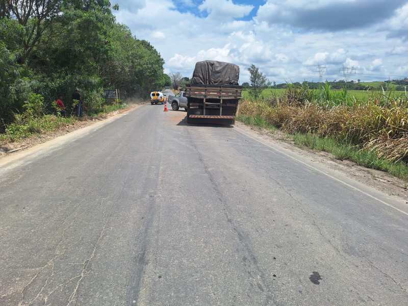 Estrada rural com um caminhão parado e um carro em movimento, cercada por vegetação e com céu limpo.