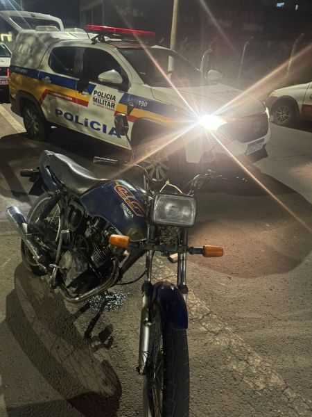 Uma motocicleta azul estacionada em uma rua, com um veículo da Polícia Militar ao fundo, iluminado por faróis.
