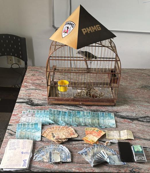 Uma gaiola com um passarinho e um copo amarelo, acompanhada de várias cédulas de dinheiro e um celular, dispostas sobre uma mesa.