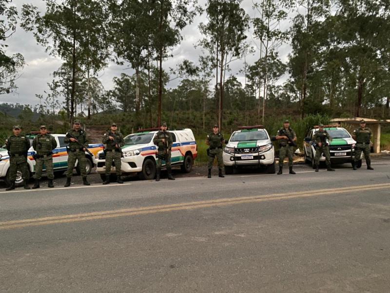 Grupo de militares armados em frente a veículos de polícia em uma estrada com árvores ao fundo.