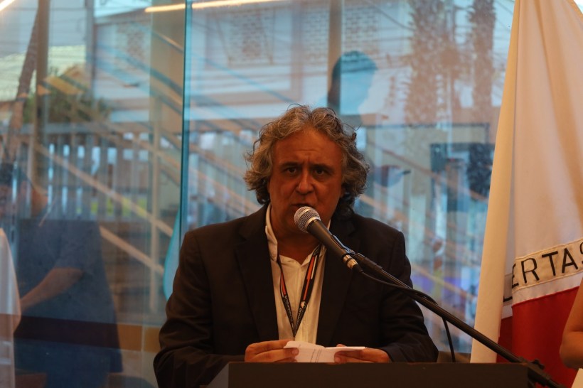 Homem com cabelo grisalho falando ao microfone em um evento, vestido com terno escuro. Ao fundo, bandeira e ambiente com iluminação natural.