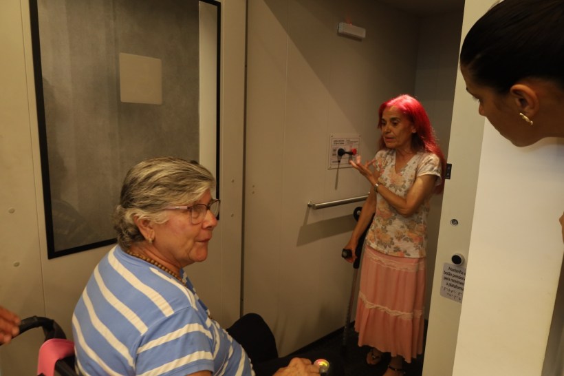 Duas mulheres conversando em um elevador, uma delas está em uma cadeira de rodas.