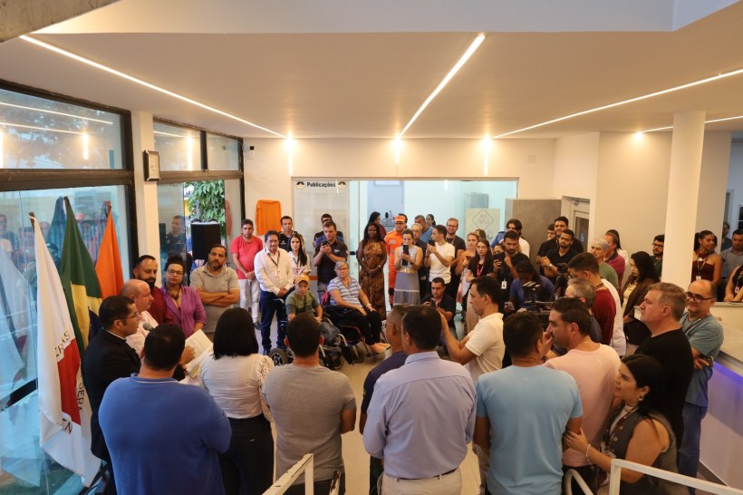 Reunião com um grande grupo de pessoas em um ambiente interno, com bandeiras ao fundo e um orador à frente, enquanto a audiência observa atentamente.