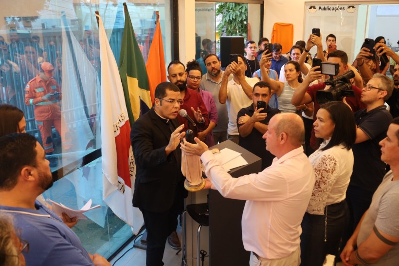 Evento de cerimônia com várias pessoas reunidas, onde um homem de terno fala ao microfone e apresenta uma estatueta a outro homem. Bandeiras ao fundo e várias pessoas filmando com celulares.