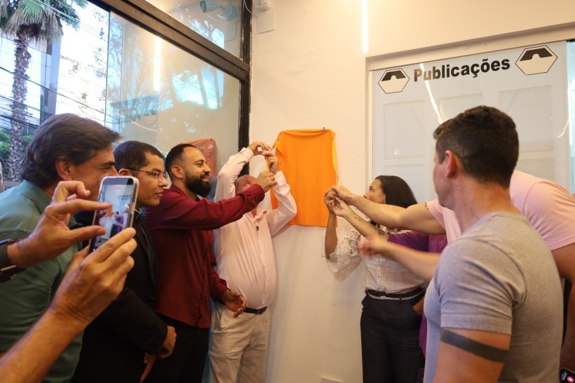 Cerimônia de descerramento de placa com várias pessoas segurando um pano laranja em um ambiente interno.