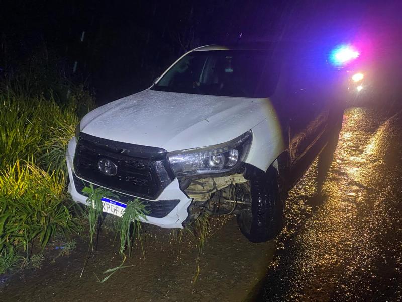 Veículo branco danificado na estrada com grama ao redor e luzes de emergência ao fundo.