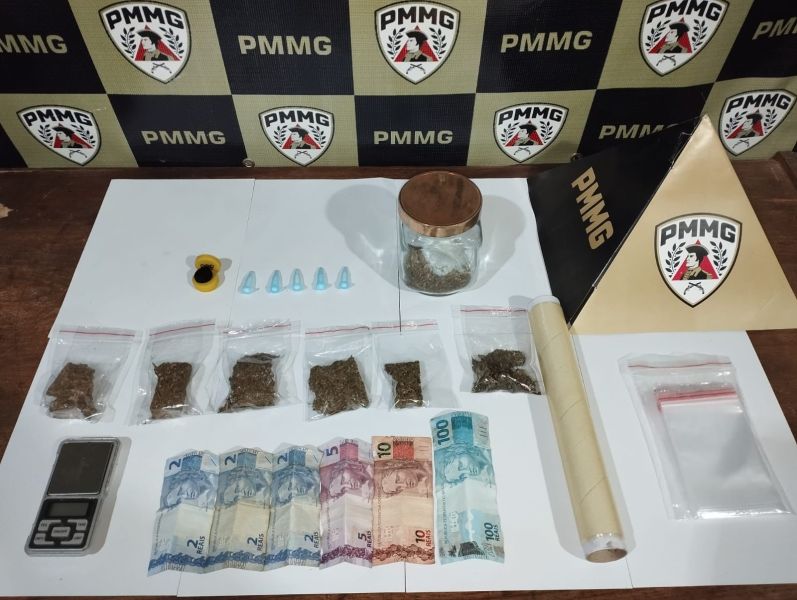 Imagem mostrando uma mesa com várias itens relacionados a substâncias ilícitas, incluindo frascos com conteúdo vegetal, pacotes plásticos, uma balança, notas de dinheiro e uma pirâmide com a logo da PMMG ao fundo.