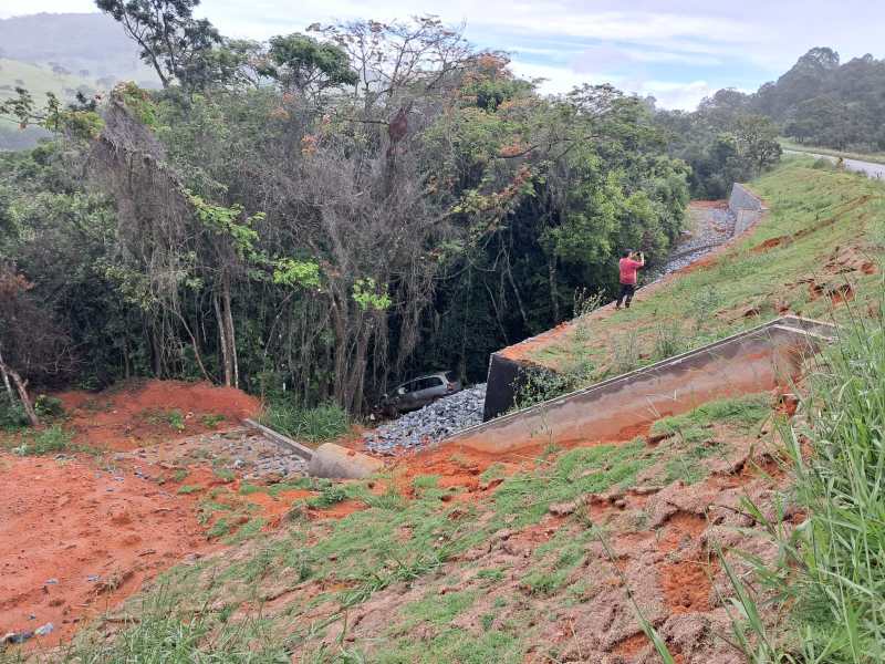 Imagem de um deslizamento de terra ao lado de uma estrada, com uma pessoa em pé tirando fotos e um carro visível na vegetação abaixo.