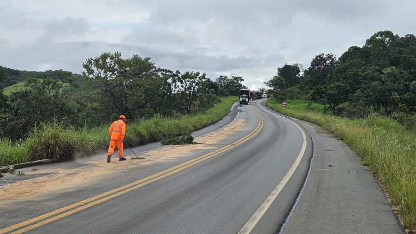Funcionário usando uniforme laranja limpando um trecho de estrada enquanto caminhões passam ao fundo.