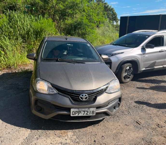 Um carro Toyota dianteiro de cor cinza estacionado em um terreno com vegetação ao fundo, ao lado de um SUV prateado.