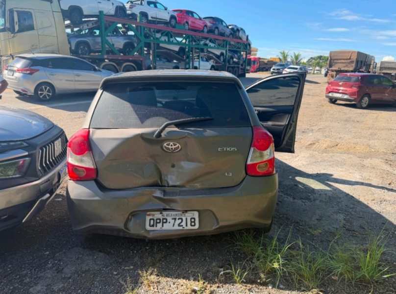 Um carro Toyota Etios cinza com a traseira danificada, estacionado em um pátio de veículos, enquanto caminhões e outros carros estão ao fundo.