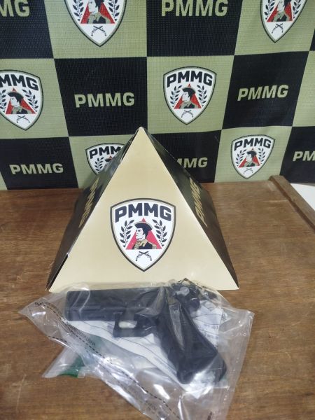 Uma arma de fogo envolta em um plástico, posicionada ao lado de uma pirâmide com o logotipo da PMMG, em um fundo quadriculado com as mesmas marcas.