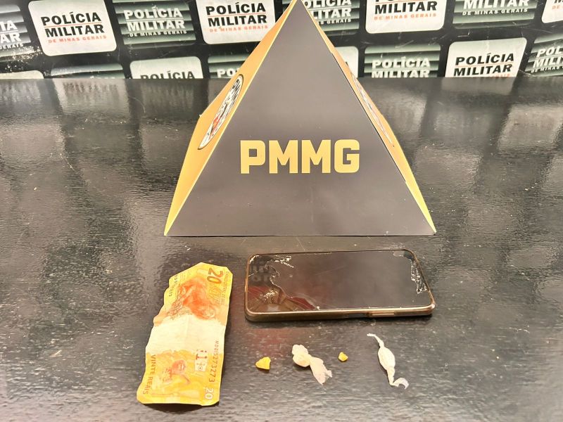 Objetos apreendidos incluindo um smartphone, uma nota de 20 reais e substâncias não identificadas sobre uma mesa com fundo da Polícia Militar.