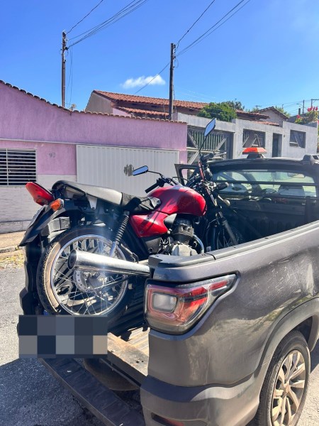 Uma moto vermelha e uma moto preta carregadas na caçamba de uma caminhonete, com o céu azul ao fundo e casas ao redor.