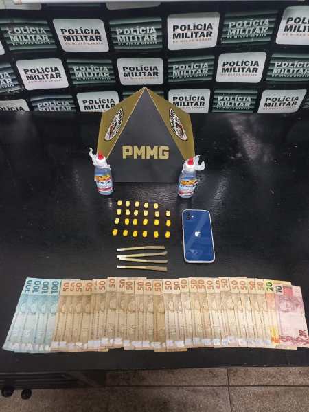 Imagem de uma mesa com dinheiro em notas brasileiras, uma faca, um celular e dois frascos. Ao fundo, painéis com a inscrição 'Polícia Militar'.