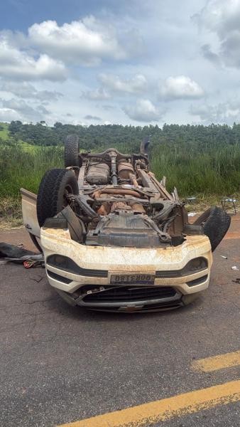 Carro capotado na estrada, visto de cima, com a parte de baixo exposta, rodeado por vegetação e céu nublado.