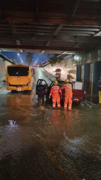 Equipes de resgate ajudam uma pessoa em um carro parcialmente submerso em água, sob um viaduto, com uma van próxima.