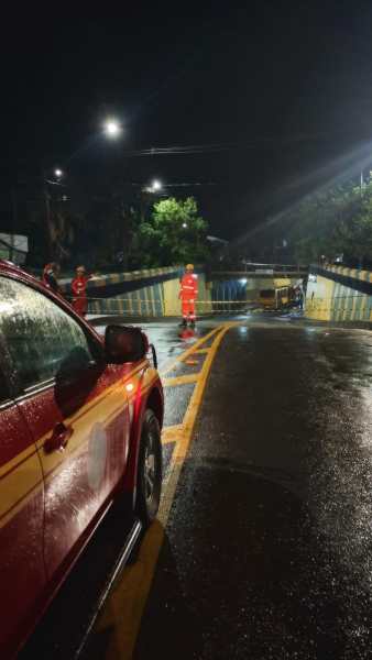 Imagem de uma estrada molhada à noite, com um carro vermelho estacionado em primeiro plano e um grupo de pessoas vestindo roupas de emergência ao fundo, próximo a uma barreira de segurança levantada.
