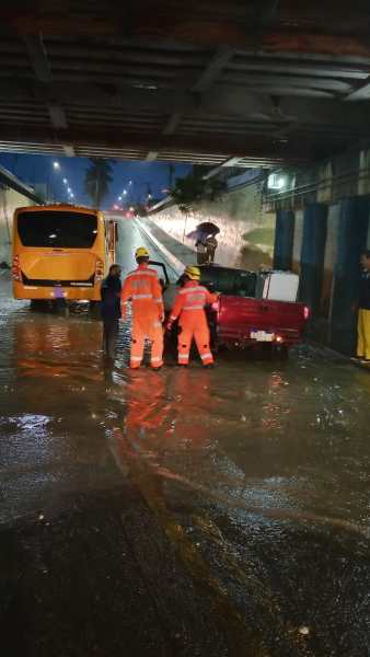 Resgate em área alagada com dois profissionais usando uniformes laranjas, um carro vermelho preso na água e um ônibus amarelo ao fundo.