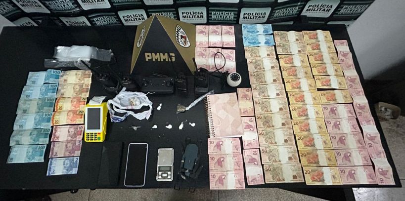 Mesa com dinheiro em notas brasileiras, incluindo cédulas de 10, 20, 50, 100 reais, aparelhos eletrônicos, drogas e materiais diversos, com fundo da polícia militar.