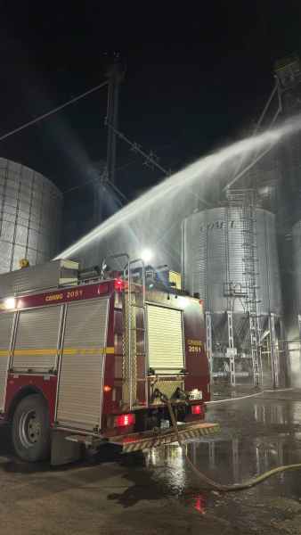 Um caminhão de bombeiros vermelho em operação durante a noite, pulverizando água em silos metálicos.