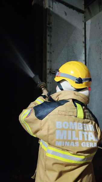 Bombeiro militar com uniforme e capacete amarelo, utilizando uma mangueira para combater um incêndio em um ambiente escuro.