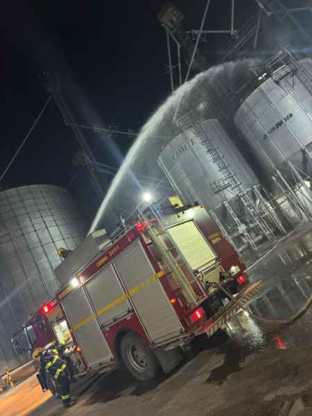 Caminhão de bombeiros em operação durante a noite, com um jato de água direcionado a silos de armazenamento metálicos.