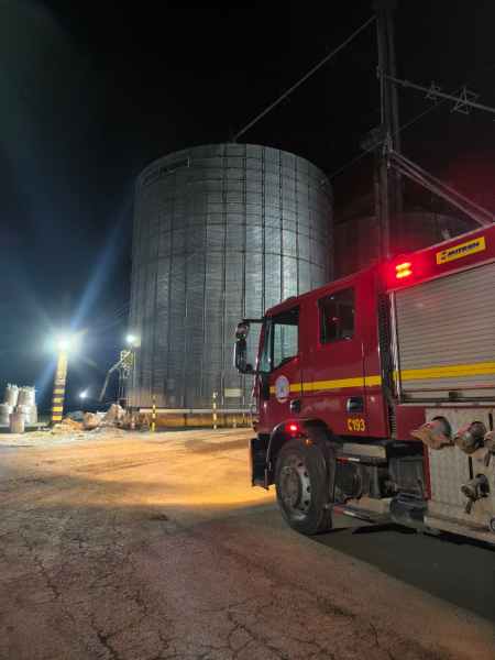 Caminhão de bombeiros em frente a um silo metálico iluminado à noite.