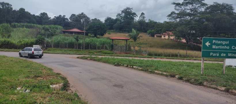 Vista de uma estrada rural com sinalização para Pitangui e Martinho Campos, cercada por vegetação e casas ao fundo.