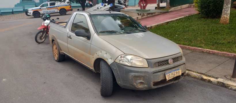 Uma caminhonete cinza suja estacionada em uma rua, com uma motocicleta e um carro da polícia ao fundo.