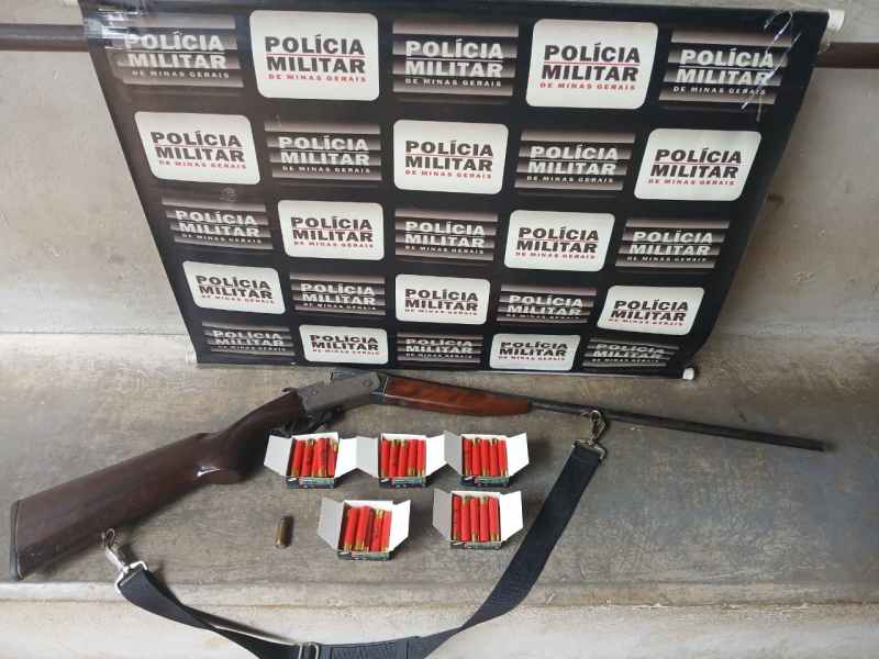 Uma espingarda de madeira, cartuchos de munição e um projétil sobre uma superfície, com um fundo de placa da Polícia Militar de Minas Gerais.