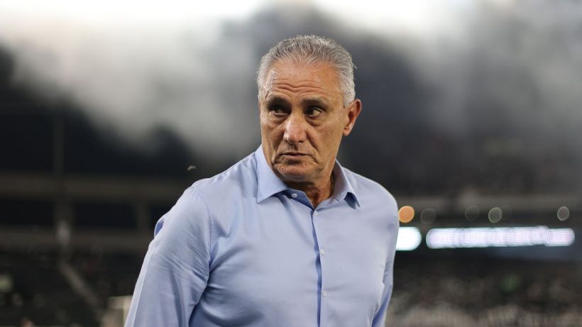 A serious-looking man with gray hair wearing a light blue shirt, standing on a sports field with smoke in the background.