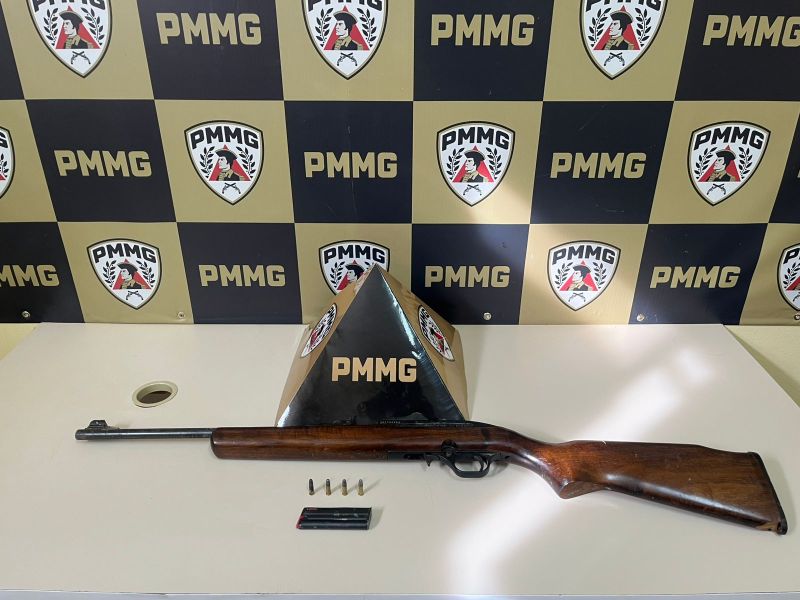 Uma espingarda com coronha de madeira e cano metálico, acompanhada de cápsulas de munição, está exibida sobre uma mesa ao lado de um suporte com o logotipo da PMMG, em um fundo com painéis pretos e dourados.