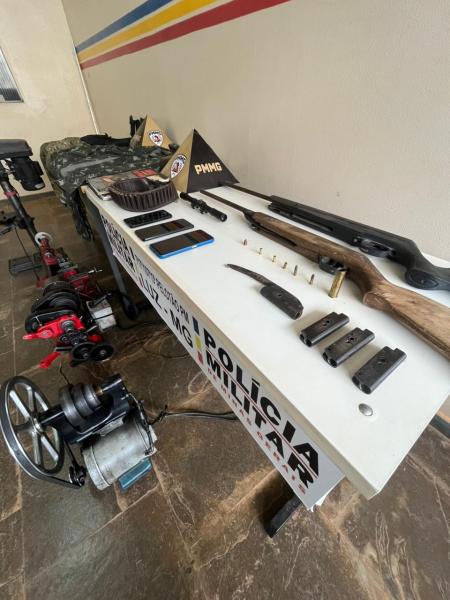 Mesa com armamentos e equipamentos da Polícia Militar, incluindo rifles, cartuchos e acessórios, em um ambiente de exibição.